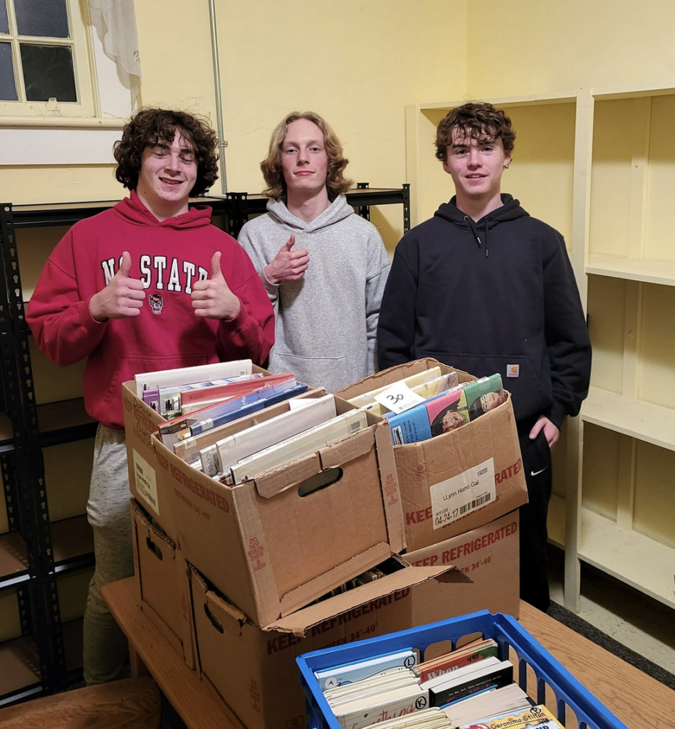 Three teenage boys volunteering at the book bank with boxes full of donated books.