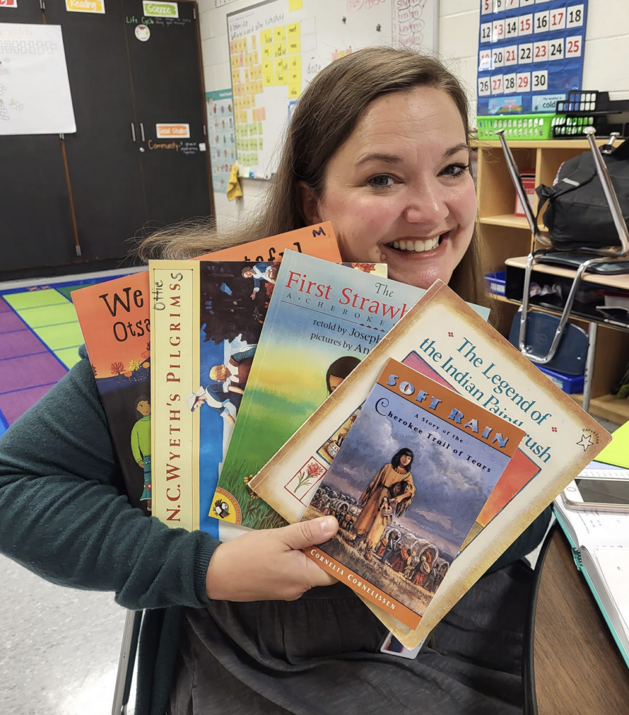 a smiling teacher standing in her haywood classroom holding up five children's books from anna's books all of them aiming to increasing representation in her classroom through diverse subject matter