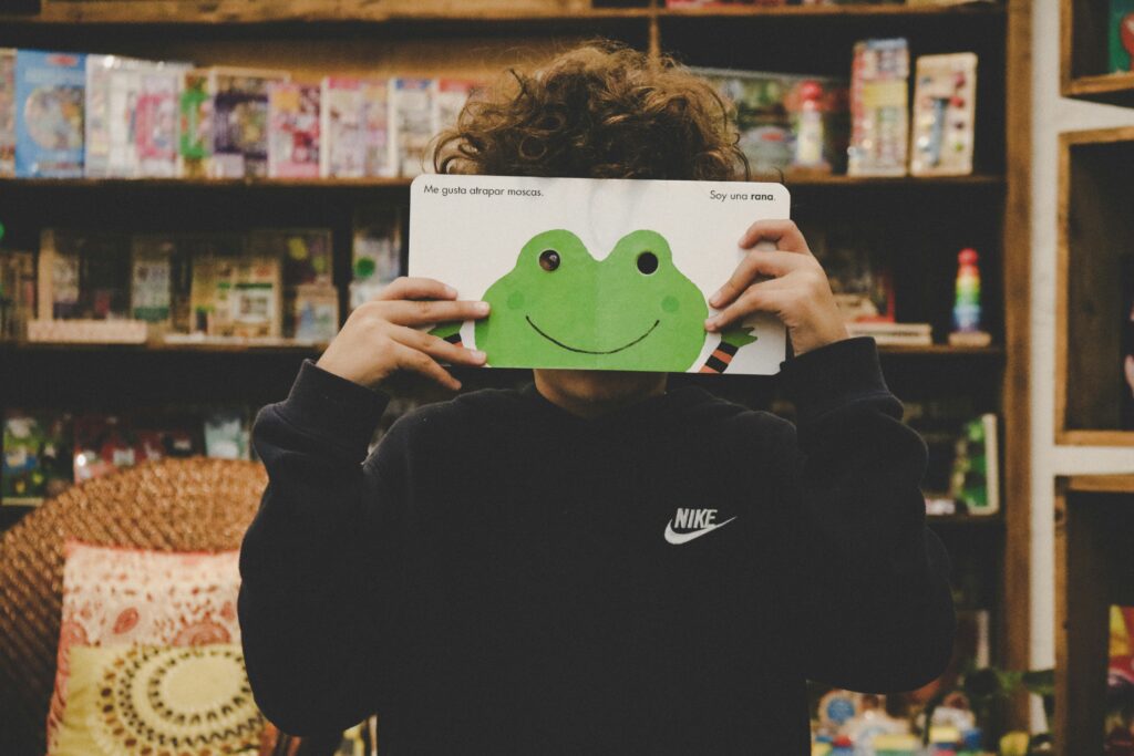 elementary school boy holding up a spanish children's book with a frog on it that covers his face except the eye holes which he peeps out of. 